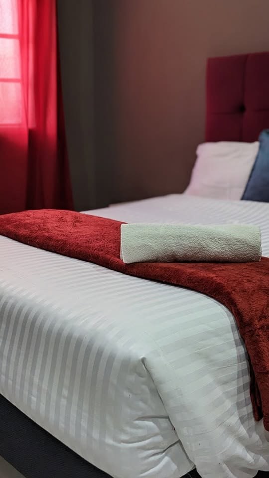 Bed with white striped linen, a red throw and a rolled towel, with red curtains at the window